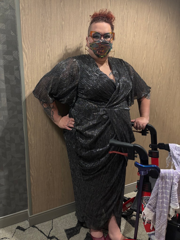 photo of a white nonbinary femme wearing glasses and a sparkly mask, standing in a sparkly dress next to their walker which has a keffiyeh draped on it