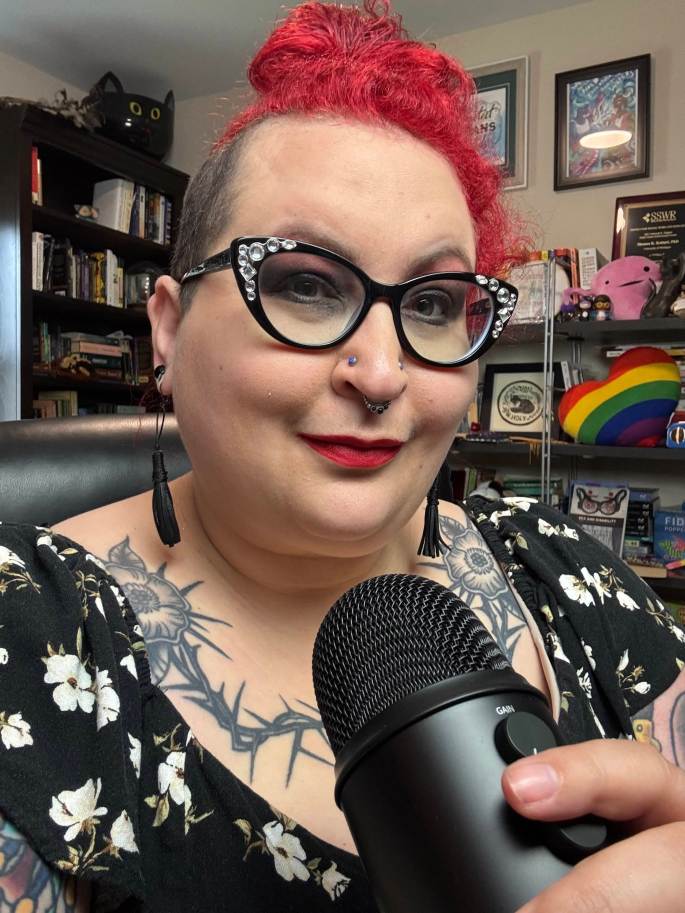 selfie of a white redheaded nonbinary femme with glasses and funky earrings with a microphone